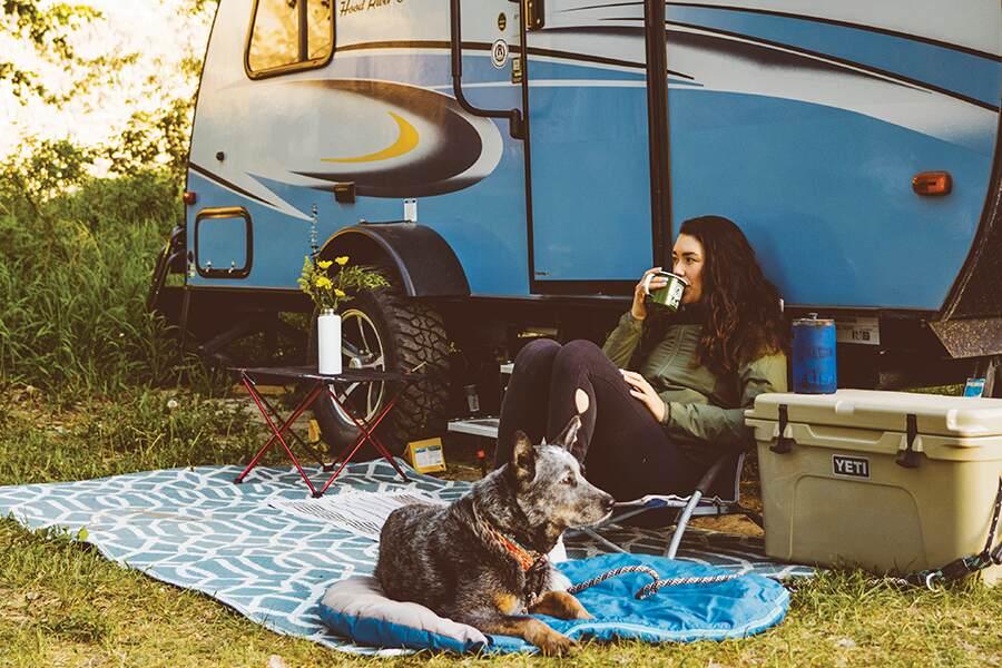 Woman relaxing with a drink beside her dog at a campsite with a travel trailer in the background.