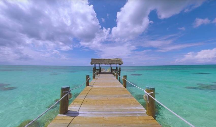 Wooden pier leading to a thatched-roof gazebo over clear turquoise waters under a partly cloudy sky.