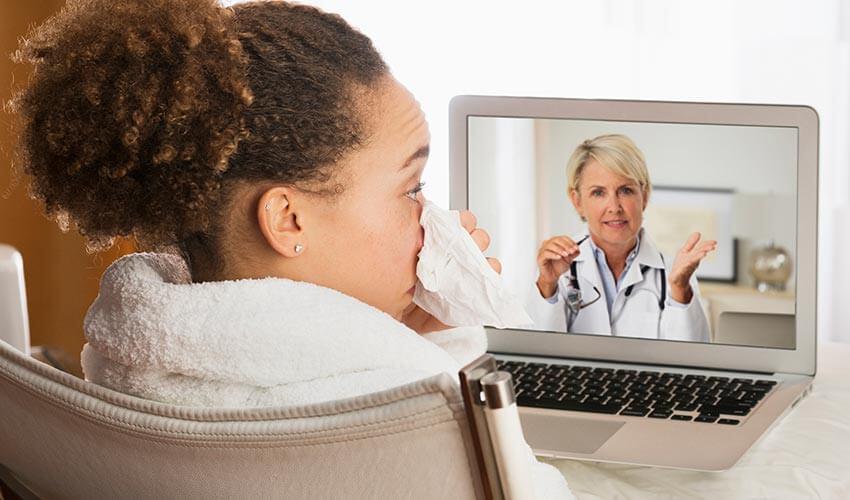 A woman is sitting in front of a laptop with a doctor on the screen.