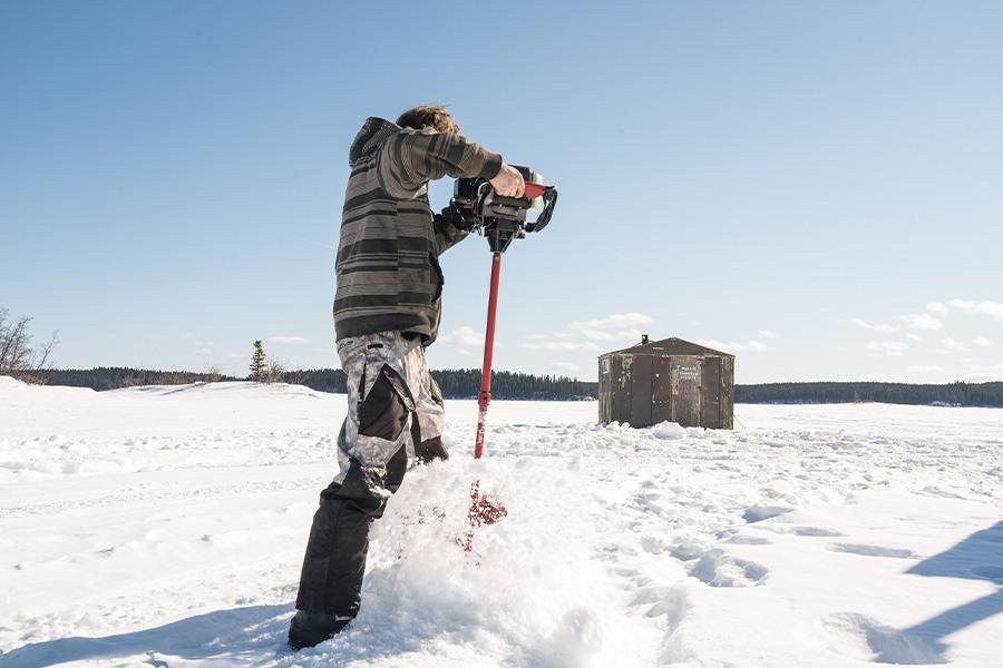Man drilling a hole in the ice for fishing near a hut on a snowy landscape.