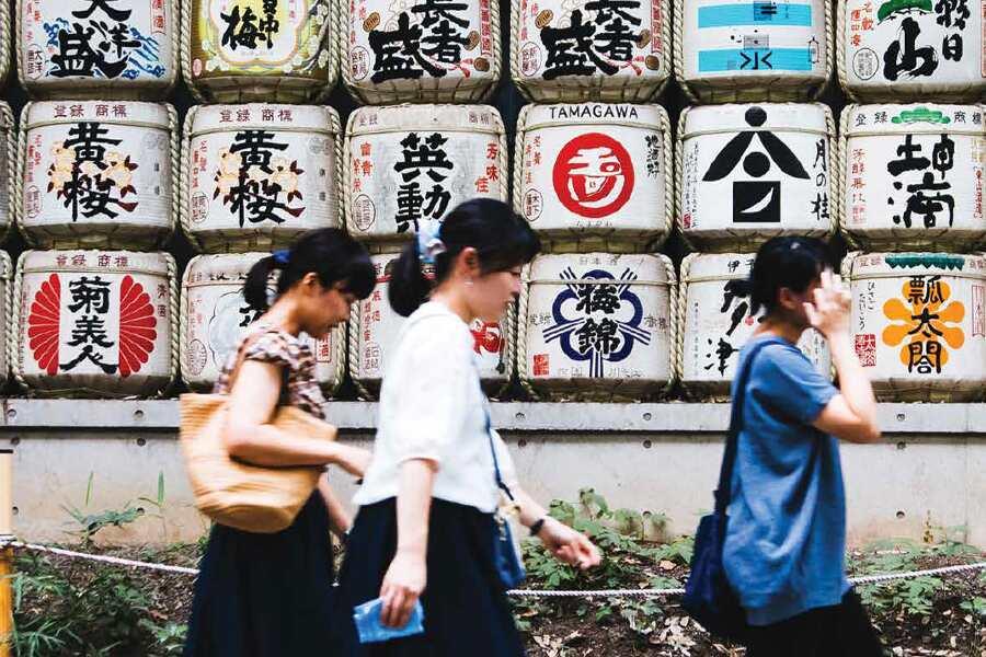 A group of women walking past a wall of barrels with japanese writing.