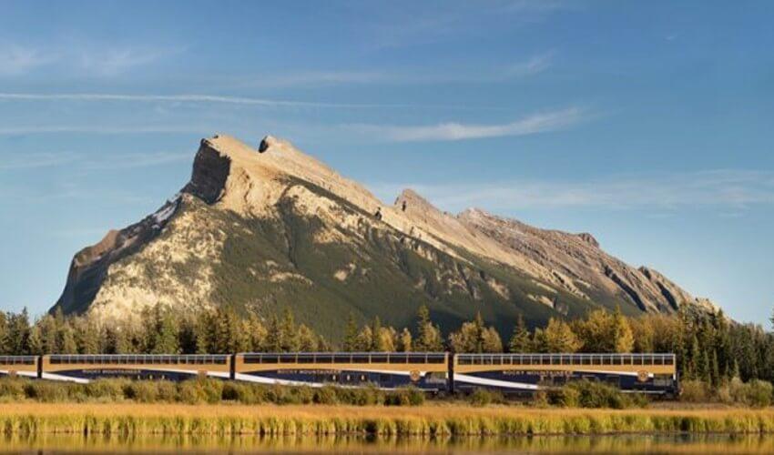 A train traveling along a river with mountains in the background.