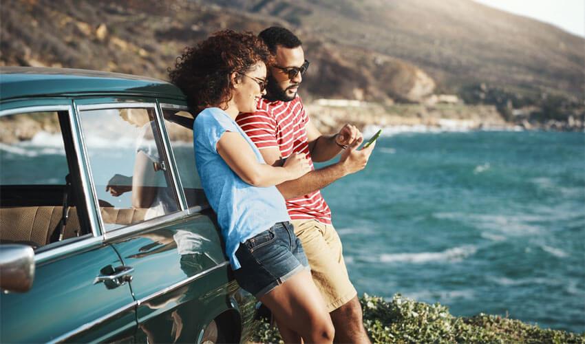 A couple looking at their phone while sitting on the side of a car by the ocean.