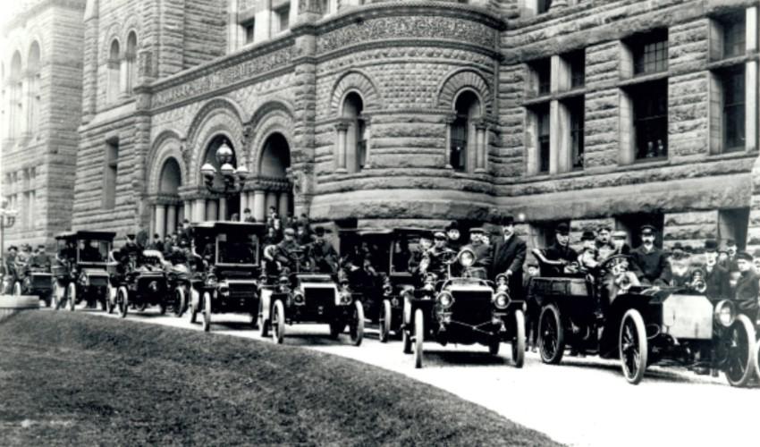 A row of early 20th-century automobiles lined up on a city street outside an ornate stone building.