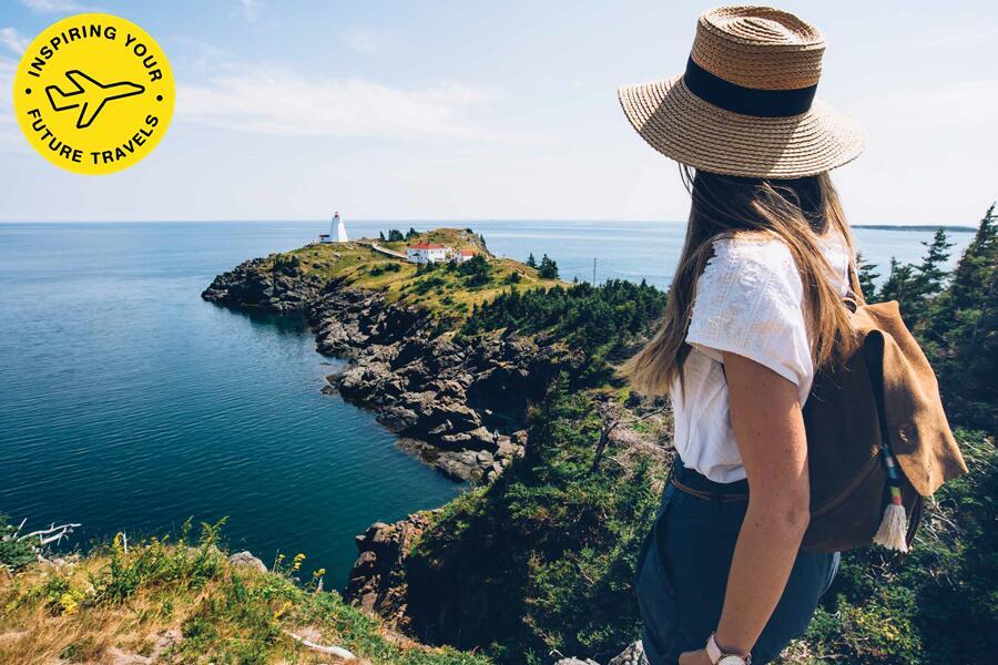 Woman in a straw hat looking out at a lighthouse on a coastal cliff.