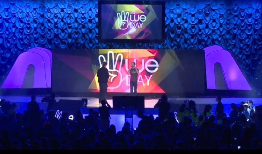 Two figures on a stage at a "we day" event with a crowd in the foreground and a lit backdrop displaying the event's logo.