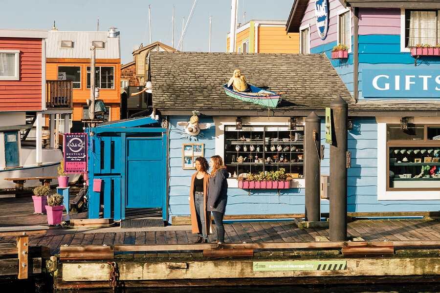 A person standing on a dock by colorful waterfront shops on a sunny day.